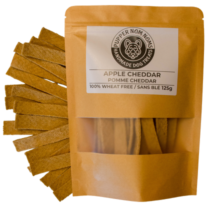 A stand-up pouch of Pupper Nom Nom Homemade Dog Treats, Apple Cheddar, shown with several rectangular cheddar treats fanned out to the left on a white background.