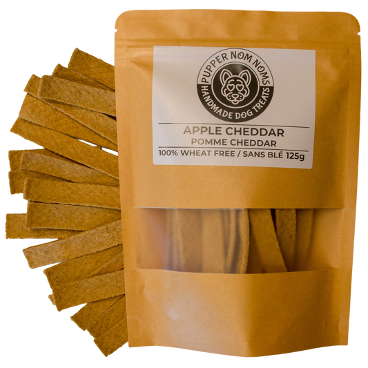 A stand-up pouch of Pupper Nom Nom Homemade Dog Treats, Apple Cheddar, shown with several rectangular cheddar treats fanned out to the left on a white background.